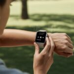 Person wearing a smartwatch with audiobook playback controls on the screen, outdoors in a park setting with wireless earbuds nearby.
