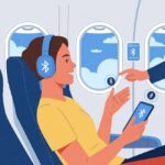 A passenger wearing Bluetooth headphones seated inside an airplane with a flight attendant nearby.