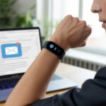 Person wearing a fitness tracker on their wrist, looking at it while sitting at a desk with a laptop showing an email inbox.