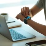 A person using a smartwatch while working on a Mac laptop at a desk with a coffee cup and notebook nearby.