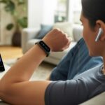 A person sitting on a couch wearing a fitness tracker and wireless earbuds, looking at the tracker while listening to a podcast, with a smartphone nearby showing a podcast app.