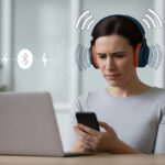 A person wearing Bluetooth headphones at a desk looks frustrated as signal waves around the headphones fade and show interference.