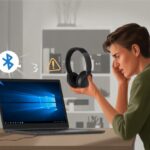 A person sitting at a desk holding Bluetooth headphones, looking confused while facing a laptop showing the Windows 10 desktop.