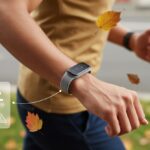 A person wearing a fitness tracker on their wrist outdoors with autumn leaves falling around them, showing the device monitoring for falls.