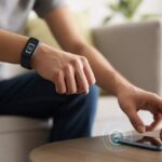 A person wearing a fitness tracker on their wrist uses it to locate a smartphone on a nearby table.