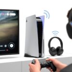 A person holding a PlayStation controller near a PlayStation console and wireless Bluetooth headphones connected to a TV displaying a game.