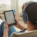 A person sitting in a living room wearing Bluetooth headphones and holding a Kindle e-reader with an open book displayed.