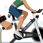 A person using a fitness tracker while riding a Peloton bike indoors, with a tablet showing workout data connected to the tracker.